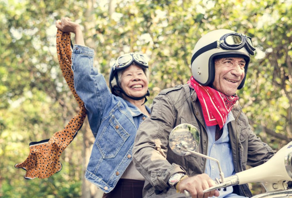 Two seniors wearing helmets and goggles enjoy a motorcycle ride through the countryside.