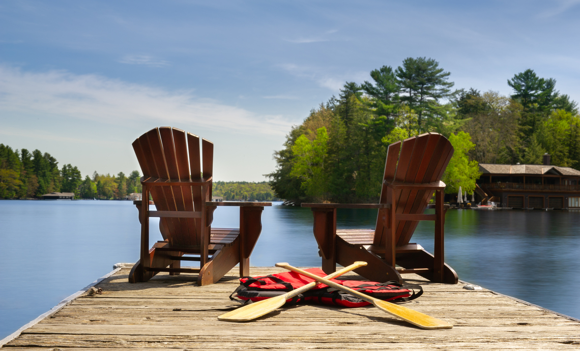 Two muskoka chairs on the end of a dock at a sunny lake in Canada.
