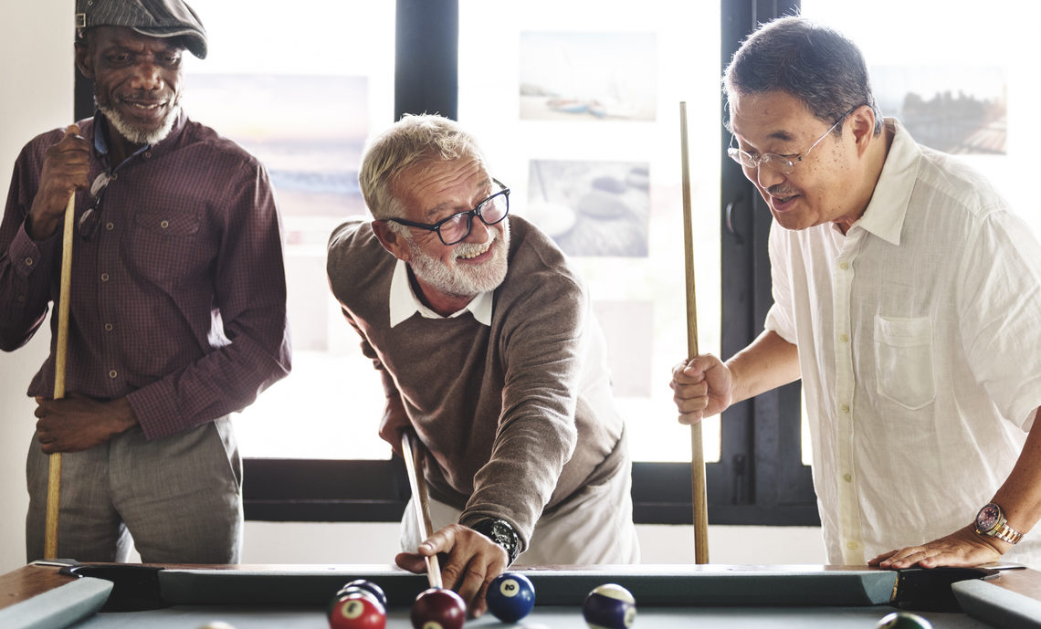 Trois retraités s’amusent et jouent au billard ensemble. L’un d’eux se prépare à jouer sous le regard des deux autres.