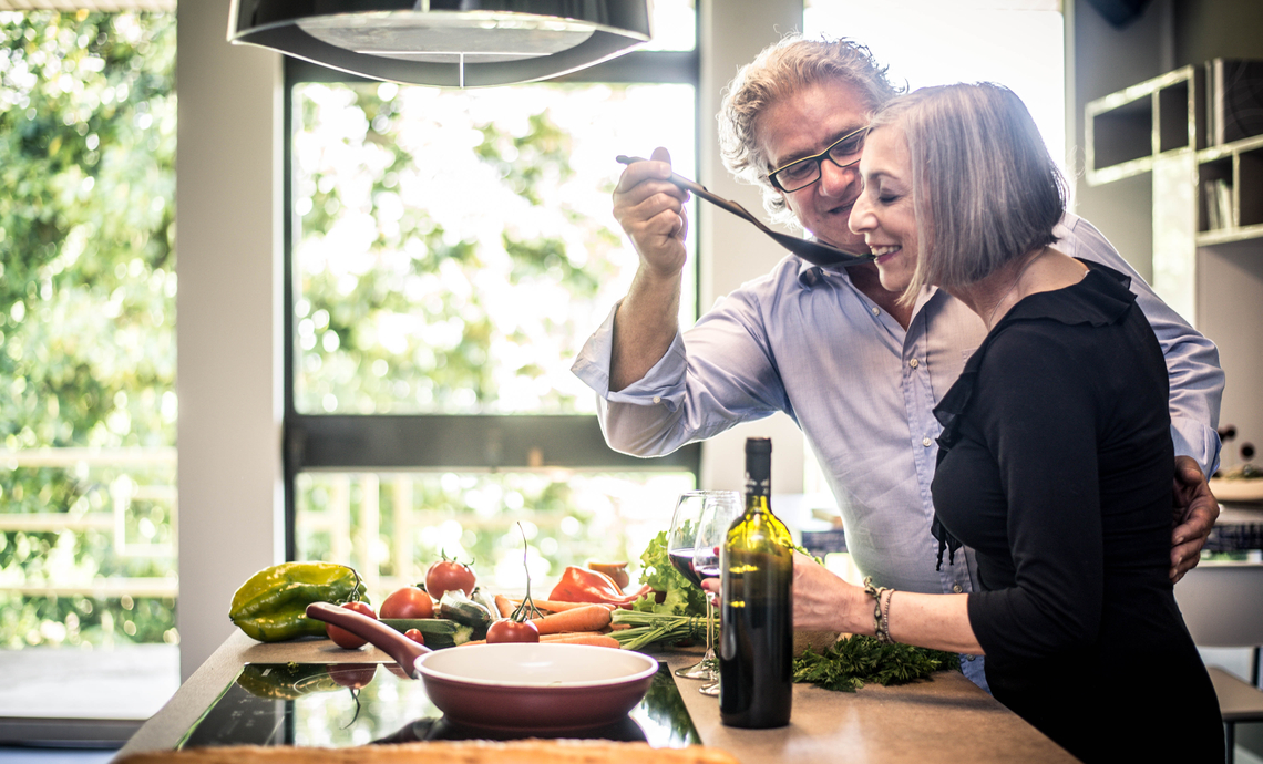 A happy couple cooking with fresh, local produce at home in their sun-filled kitchen.