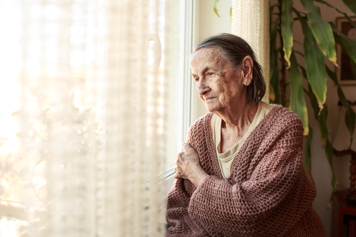 Une aînée au regard triste vivant dans un établissement de soins de longue durée regarde à l'extérieur, appuyée contre une fenêtre.