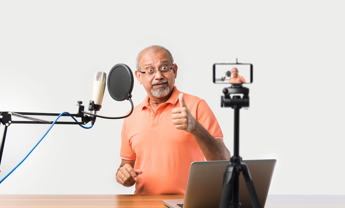 Retiree standing in front of a laptop, with audio and video equipment set up recording a podcast and video.