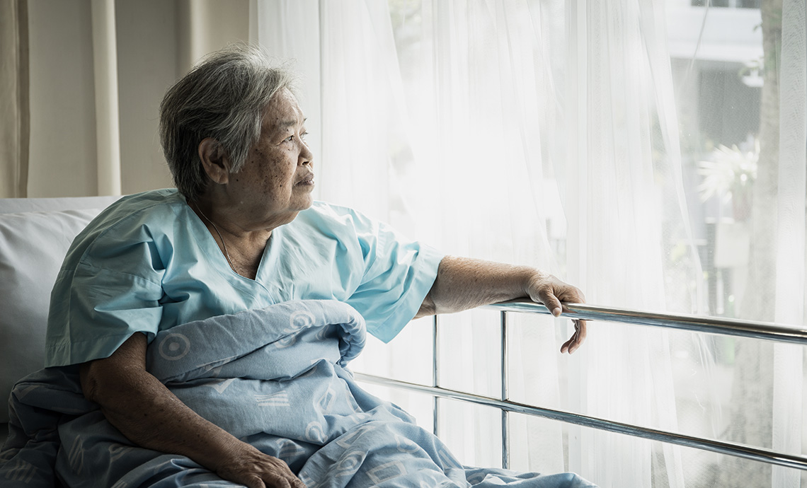 une personne âgée assise dans son lit dans une maison de soins de longue durée et regardant par la fenêtre, semblant se sentir seule